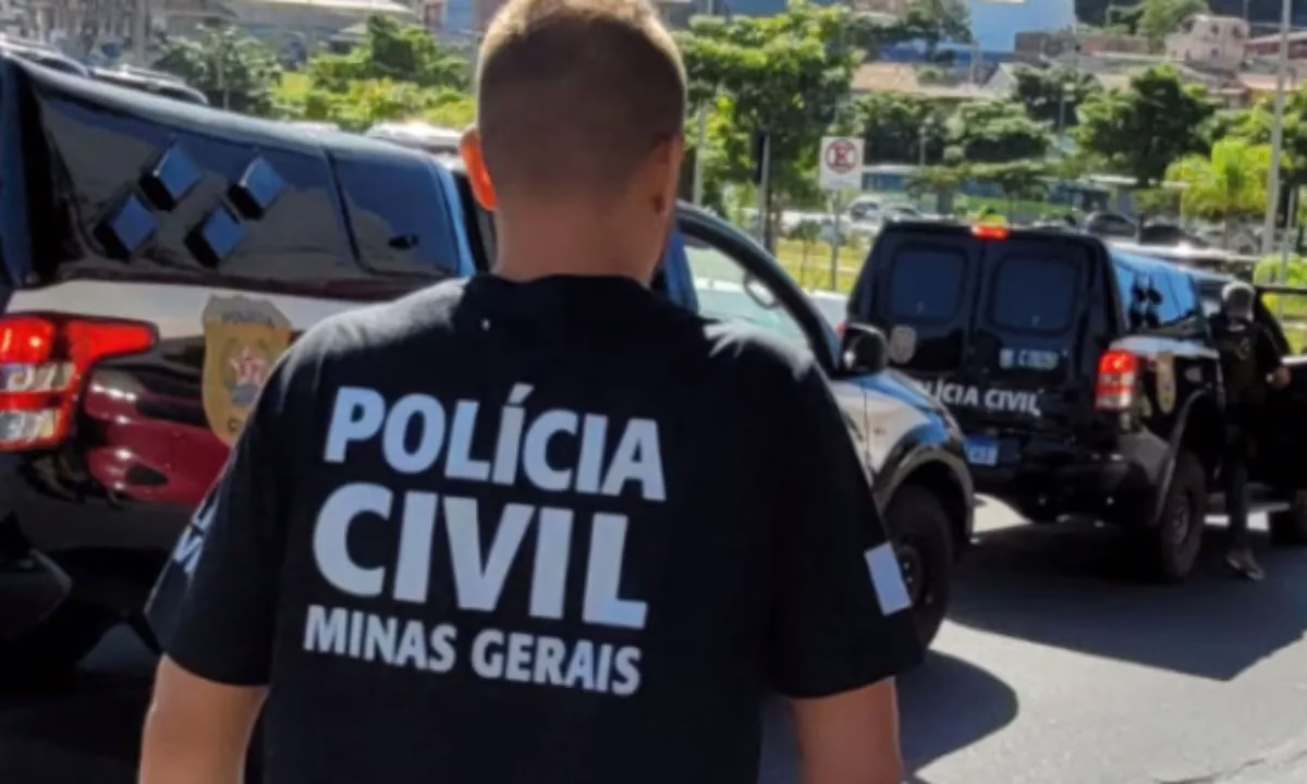 Policial civil de Minas Gerais, visto de costas com camiseta da Polícia Civil, diante de viaturas da corporação em operação.
